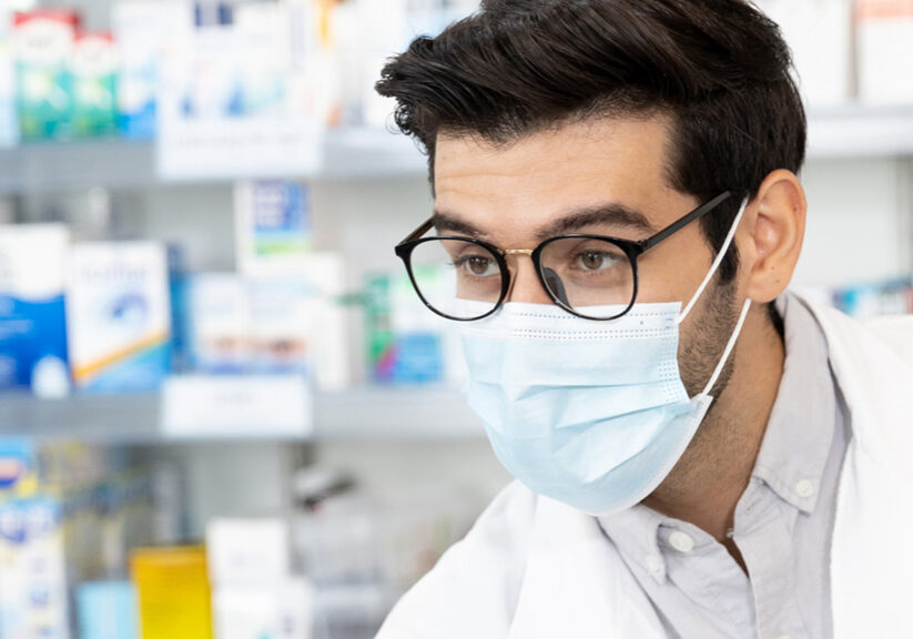 Middle eastern male pharmacist wearing protective hygienic mask to prevent infection selling medications to woman patient to prescription and making drug recommendations in modern pharmacy